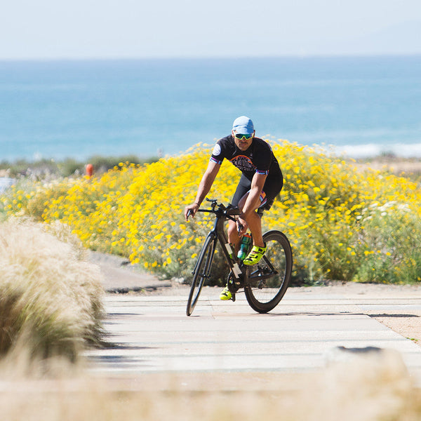 Man on bike near ocean wearing blue 4-panel cap.